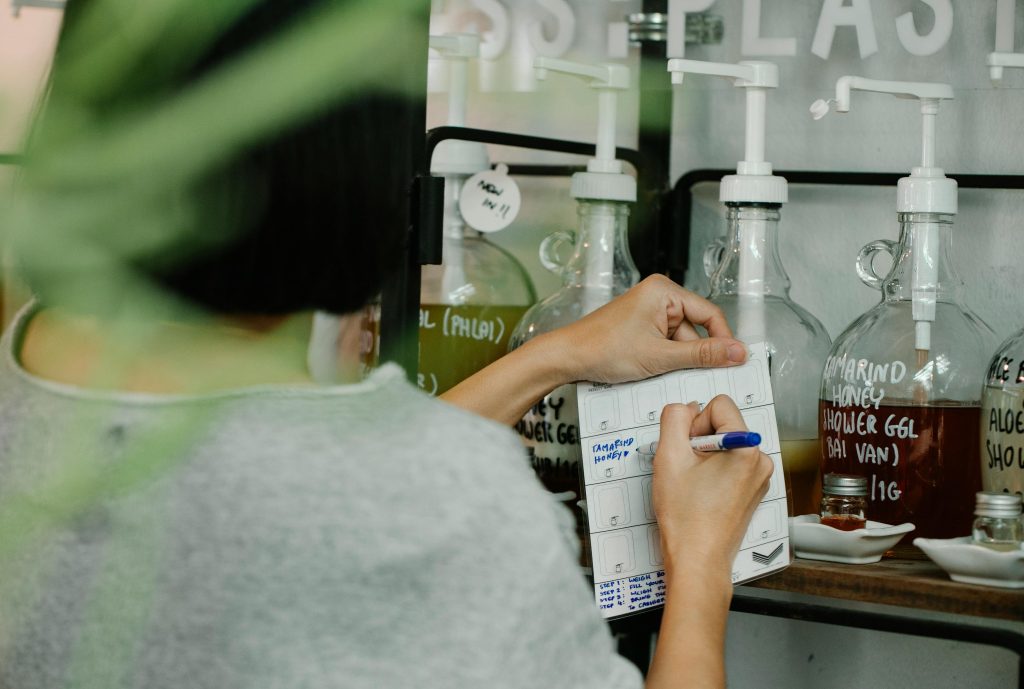 pexels-photo-7262690-7262690 Person using a pen to take notes in an eco-friendly refill store with glass containers.